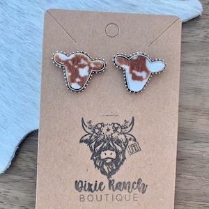 May include: A pair of cowhide earrings with silver accents. The earrings are displayed on a brown card with a cow logo and the text "Dixie Ranch Boutique".