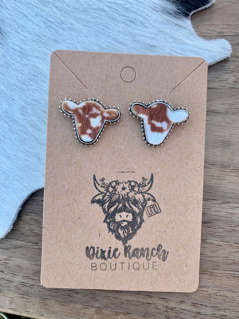 May include: A pair of cowhide earrings with silver accents. The earrings are displayed on a brown card with a cow logo and the text "Dixie Ranch Boutique".