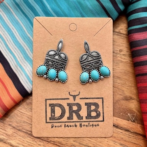 May include: A pair of silver-toned earrings with turquoise-colored stones. The earrings are on a brown card with the text "DRB Dixie Ranch Boutique". The background features a colorful fabric with stripes and patterns.
