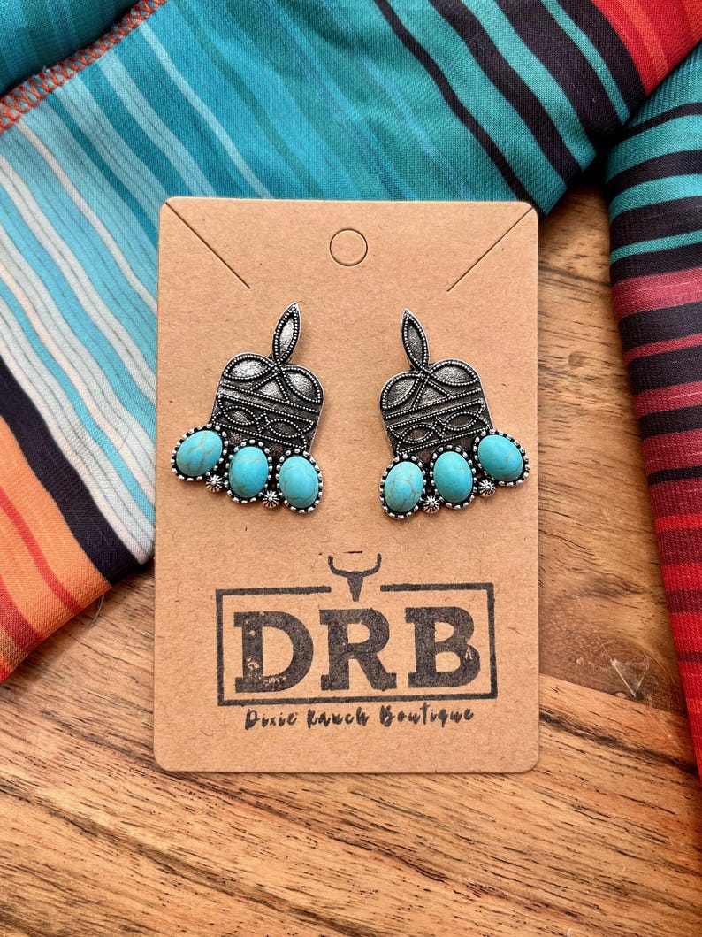 May include: A pair of silver-toned earrings with turquoise-colored stones. The earrings are on a brown card with the text "DRB Dixie Ranch Boutique". The background features a colorful fabric with stripes and patterns.