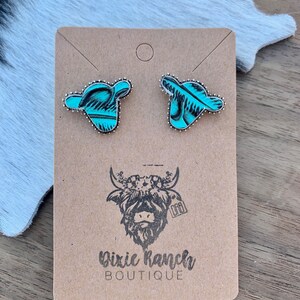 May include: A pair of turquoise and silver cow head stud earrings on a brown card with the text "Dixie Ranch Boutique" and a black and white illustration of a cow with a floral crown.