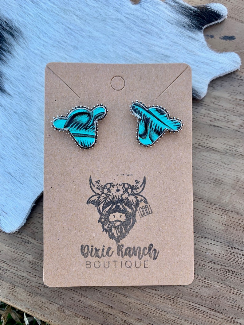 May include: A pair of turquoise and silver cow head stud earrings on a brown card with the text "Dixie Ranch Boutique" and a black and white illustration of a cow with a floral crown.