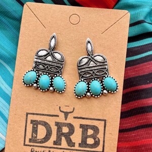 May include: A pair of silver-toned earrings with turquoise-colored stones. The earrings are on a brown card with the text "DRB Dixie Ranch Boutique". The background features a colorful striped fabric.