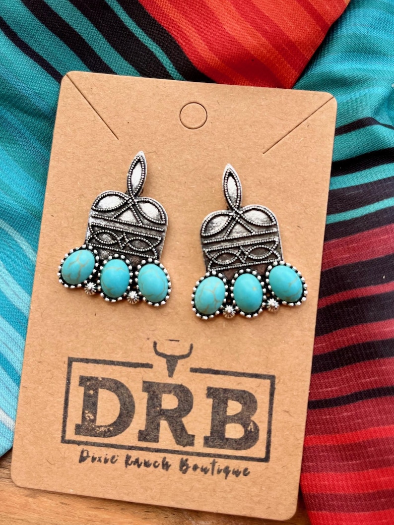 May include: A pair of silver-toned earrings with turquoise-colored stones. The earrings are on a brown card with the text "DRB Dixie Ranch Boutique". The background features a colorful striped fabric.