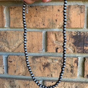 Pu&ograve; includere: Una collana di perline d'argento con una catena e una chiusura delicate. La collana &egrave; composta da piccole perline rotonde d'argento. La collana &egrave; mostrata su uno sfondo di muro di mattoni.