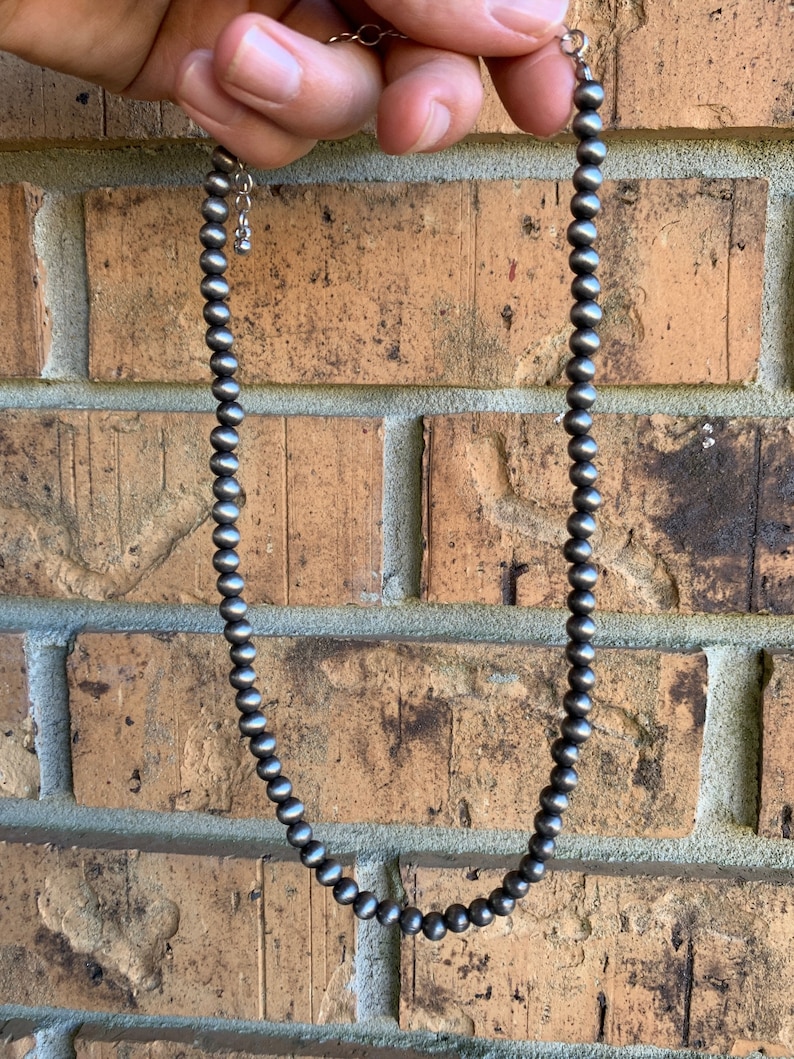 Pu&ograve; includere: Una collana di perline d'argento con una catena e una chiusura delicate. La collana &egrave; composta da piccole perline rotonde d'argento. La collana &egrave; mostrata su uno sfondo di muro di mattoni.
