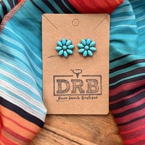 May include: Turquoise flower stud earrings on a brown card with the text "DRB Dixie Ranch Boutique". The earrings are displayed on a colourful striped fabric with shades of blue, orange, and red.