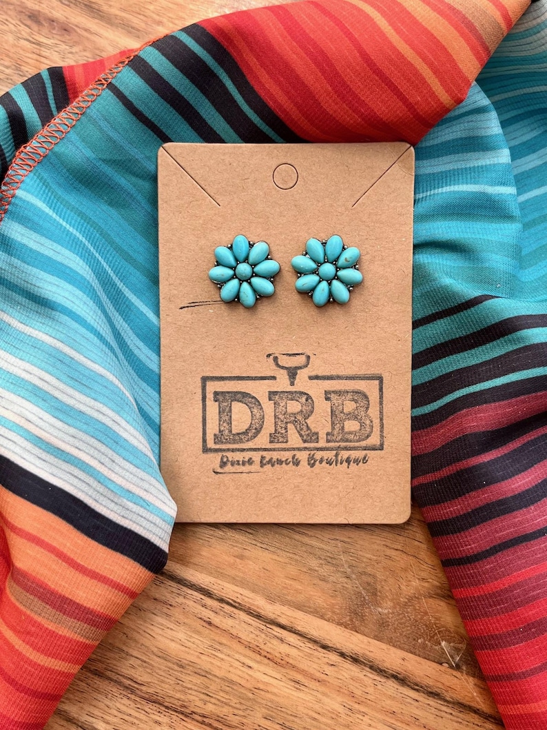 May include: Turquoise flower stud earrings on a brown card with the text "DRB Dixie Ranch Boutique". The earrings are displayed on a colourful striped fabric with shades of blue, orange, and red.