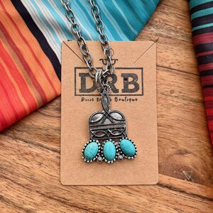 Puede incluir: Collar plateado con un colgante con tres piedras turquesas ovaladas. El colgante tiene un dise&ntilde;o ornamentado y est&aacute; unido a una cadena gruesa. El collar se exhibe en una tarjeta marr&oacute;n con el texto "DRB Pure Western Boutique".