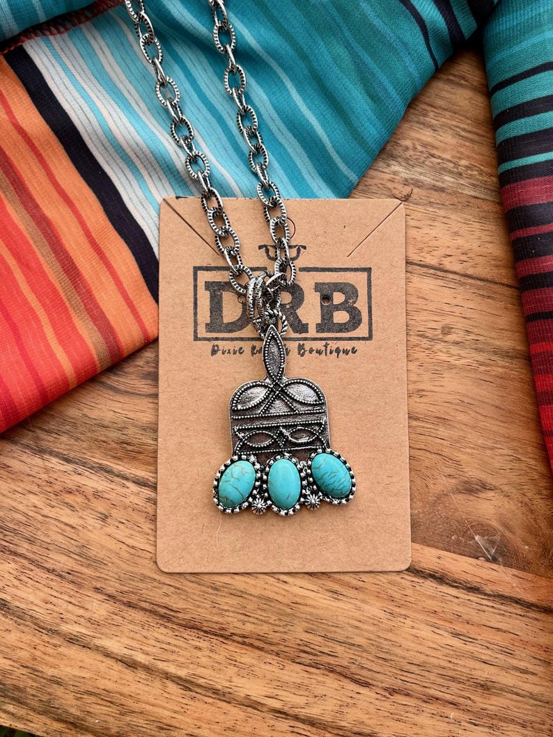 Puede incluir: Collar plateado con un colgante con tres piedras turquesas ovaladas. El colgante tiene un dise&ntilde;o ornamentado y est&aacute; unido a una cadena gruesa. El collar se exhibe en una tarjeta marr&oacute;n con el texto "DRB Pure Western Boutique".