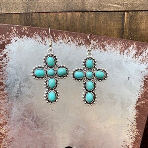 May include: A pair of silver-toned cross-shaped earrings. Each earring features turquoise-colored oval stones set within a detailed silver frame. The earrings have a hook closure for pierced ears.