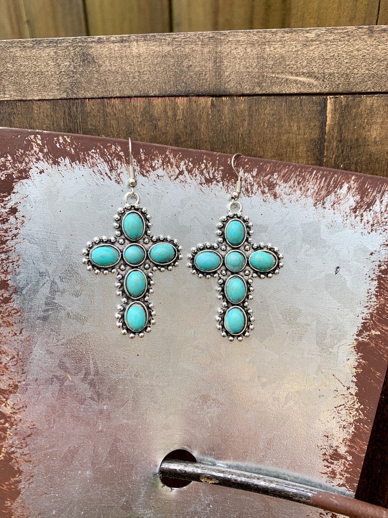 May include: A pair of silver-toned cross-shaped earrings. Each earring features turquoise-colored oval stones set within a detailed silver frame. The earrings have a hook closure for pierced ears.
