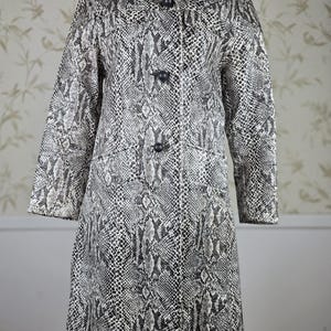 May include: A long-sleeved coat featuring a black and white snakeskin pattern. The coat has a wide collar, a button-down front with three dark circular buttons, and a straight hemline. The garment is displayed on a mannequin to show its structure.