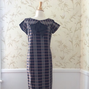 May include: A black and brown plaid dress with a large black bow at the neckline. The dress has short sleeves and a fitted silhouette.