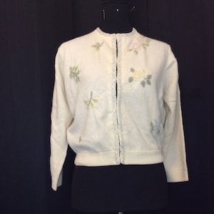 May include: A white cardigan sweater with floral embroidery. The cardigan has a pearl button closure and long sleeves.