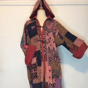 May include: A long, patchwork quilted coat with a hood. The coat is made of various fabrics in shades of pink, brown, blue, and black. The coat has a front closure with wooden toggles.