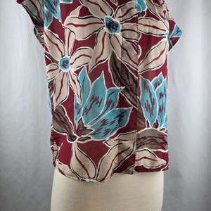 May include: A short-sleeved blouse with a floral pattern. The blouse features large flowers in shades of burgundy, beige, teal, and brown. The fabric appears to be lightweight and semi-sheer, with a button-down front and a collared neckline.