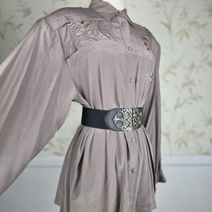 May include: A taupe long-sleeve button-down shirt featuring decorative embroidery on the upper chest panels. The shirt is cinched at the waist with a wide black elastic belt that has a large, ornate silver-toned metal buckle with intricate detailing.