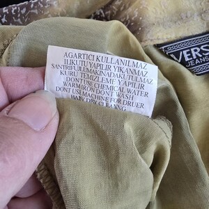 May include: Close-up of a garment with a tag featuring care instructions in multiple languages. The fabric is a muted olive green with a textured pattern. A black label with white text reading "VERS JEANS" is visible.