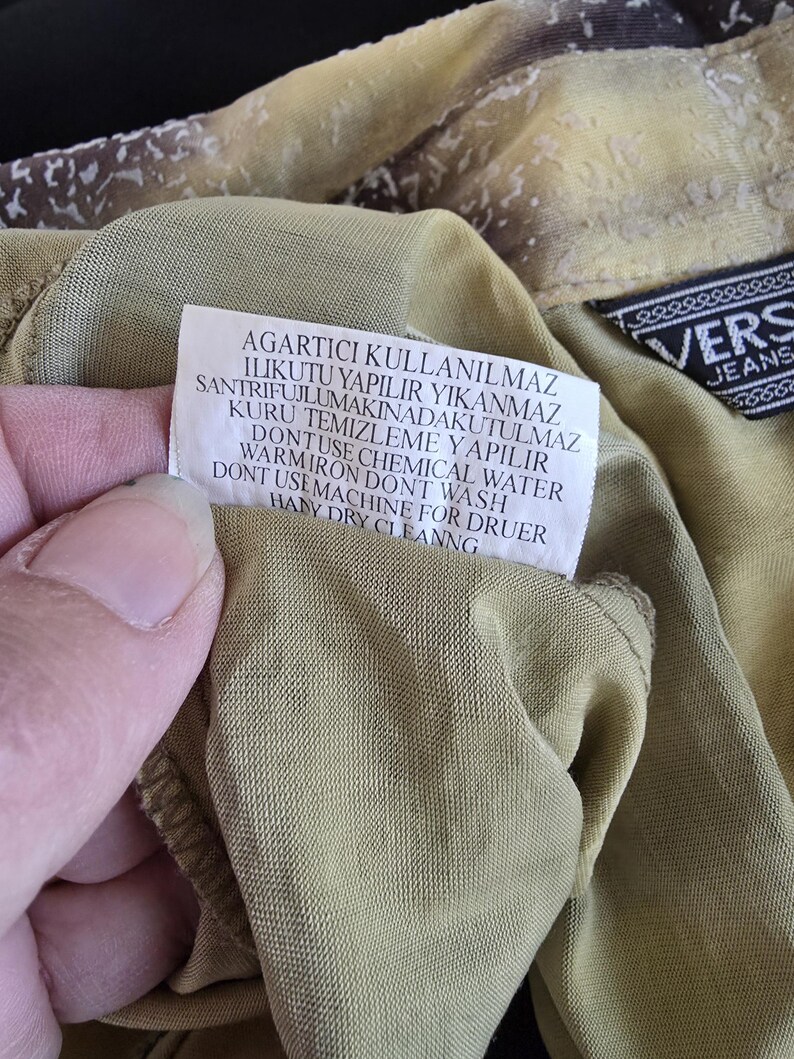 May include: Close-up of a garment with a tag featuring care instructions in multiple languages. The fabric is a muted olive green with a textured pattern. A black label with white text reading "VERS JEANS" is visible.