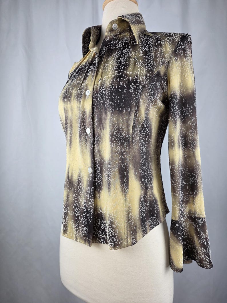 May include: A long-sleeved button-down shirt with a unique pattern. The shirt features a collared neckline and a pattern of yellow, brown, and white speckles. The sleeves have a flared design. The shirt is displayed on a mannequin.