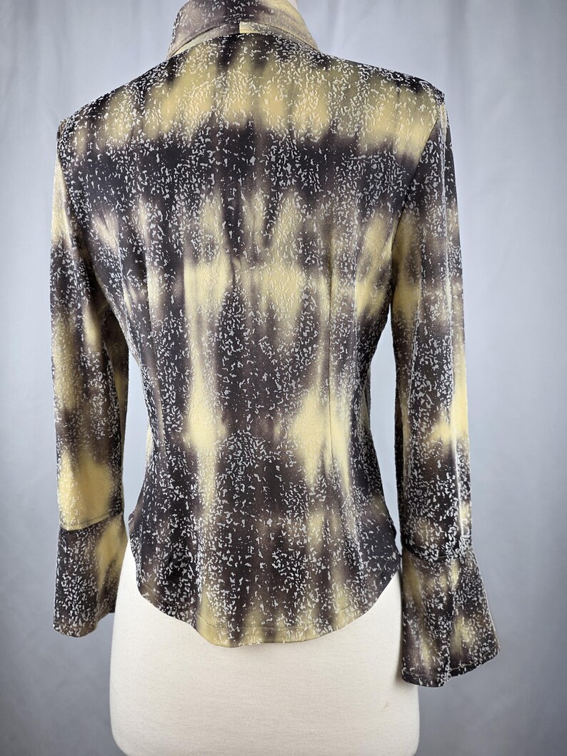 May include: A long-sleeved blouse with a unique pattern. The blouse features a collared neckline and flared sleeves. The fabric has a mottled design in shades of brown, tan, and white, creating a textured appearance. The blouse is displayed on a mannequin.