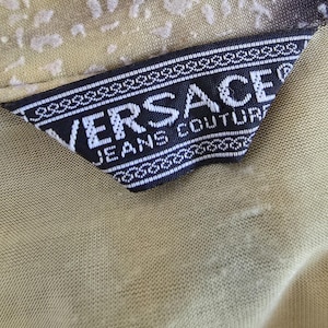 May include: Close-up of a Versace Jeans Couture label. The label is black with white text and a decorative border. The background fabric is a light olive green color with a subtle pattern.