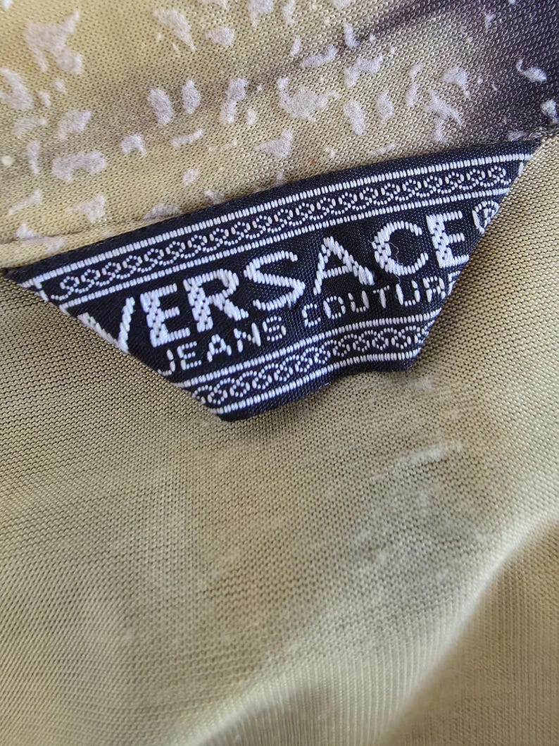 May include: Close-up of a Versace Jeans Couture label. The label is black with white text and a decorative border. The background fabric is a light olive green color with a subtle pattern.