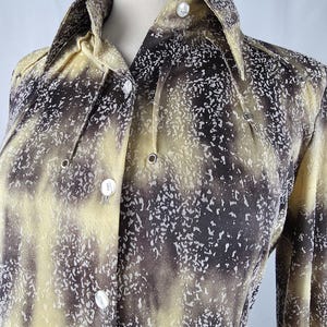 May include: A button-down shirt with a unique pattern in shades of yellow, brown, and white. The long-sleeved shirt features a collared neckline and white buttons. The fabric has a speckled design.