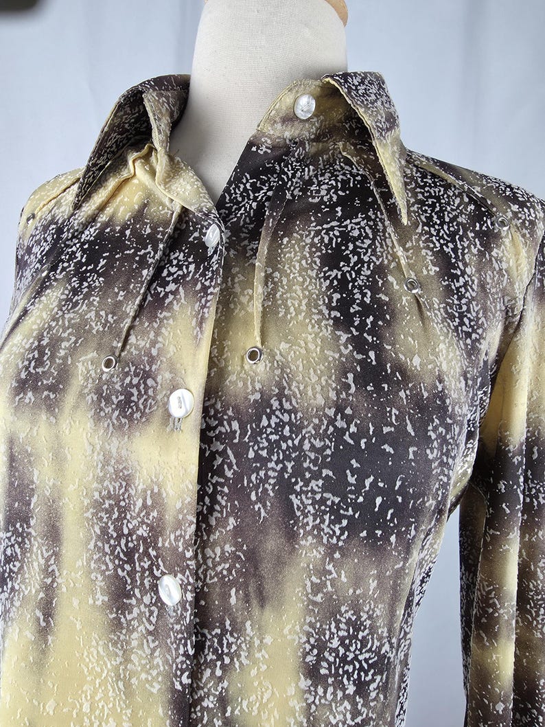 May include: A button-down shirt with a unique pattern in shades of yellow, brown, and white. The long-sleeved shirt features a collared neckline and white buttons. The fabric has a speckled design.