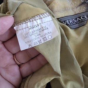 May include: Close-up of a tan-colored garment with a label that reads "IOMAN 2002 SPRING SUMMER." The label also indicates the fabric composition: 96% cotton and 4% lycra. A Versace Jeans Couture label is also visible.