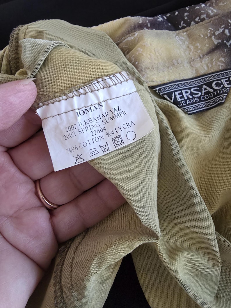 May include: Close-up of a tan-colored garment with a label that reads "IOMAN 2002 SPRING SUMMER." The label also indicates the fabric composition: 96% cotton and 4% lycra. A Versace Jeans Couture label is also visible.