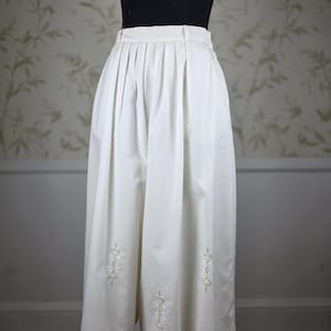 May include: White culottes with pleats and embroidered floral designs. The high-waisted pants are knee-length and feature decorative embroidery near the hem. The garment is displayed on a mannequin against a floral wallpaper background.