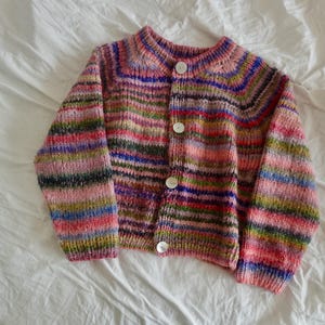 May include: A multi-coloured, hand-knitted cardigan with a round neckline and three white buttons. The sweater features horizontal stripes in shades of pink, blue, green, and purple. The garment is displayed on a white surface.