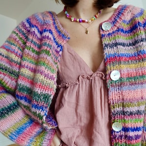 Könnte beinhalten: Ein mehrfarbiger gestreifter Strickcardigan mit großen, perlmuttartigen Knöpfen. Der Pullover hat eine Mischung aus Rosa-, Blau-, Grün- und Gelbtönen. Getragen über einem rosa Oberteil und kombiniert mit einer Jeans, entsteht ein lebendiger, vielschichtiger Look.