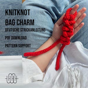 Könnte beinhalten: Ein leuchtend roter gestrickter Taschenanhänger. Der Anhänger wird in der Hand einer Person gehalten. Das Bild enthält den Text "KNITKNOT BAG CHARM", "DEUTSCHE STRICKANLEITUNG", "PDF DOWNLOAD" und "PATTERN SUPPORT".