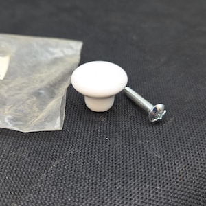 May include: A white cabinet knob with a mushroom-shaped design. The knob is accompanied by a silver screw for installation. The hardware is likely for furniture or cabinetry. The knob is approximately 1.5 inches in diameter.