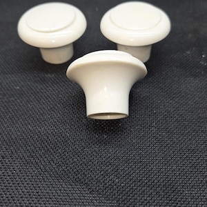 May include: Three off-white cabinet knobs with a smooth, rounded top surface and a tapered base. Each knob features a circular design with a slightly raised center section and a flared edge, providing a simple, functional hardware component.