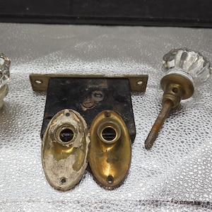 May include: An assortment of antique door hardware, including two clear glass doorknobs, a black metal lock, and two brass escutcheons. The doorknobs have a faceted design. The hardware is likely from a vintage door.