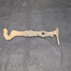 May include: A vintage bronze-colored metal key with an ornate design. The key features a curved handle, a decorative leafy section, a circular hole, and a rectangular end. The key is likely for a cabinet or small lock.