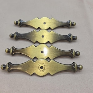 May include: Four antique brass drawer pulls with a decorative, symmetrical design. Each pull features a central diamond shape with a hole for mounting and rounded ends. The pulls have a dark, aged finish, suggesting vintage or rustic decor.