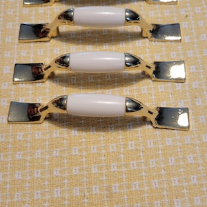 May include: Four white ceramic cabinet knobs with gold metal bases. The knobs are arranged in a row on a yellow and white patterned surface.