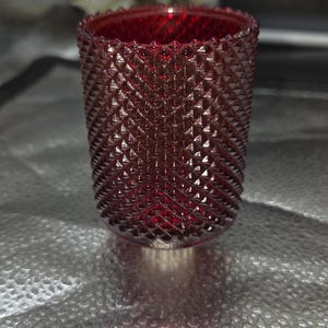 May include: A ruby red glass votive candle holder with a textured, diamond-patterned exterior. The candle holder is cylindrical with a slightly flared top. The interior is visible, and the glass appears to be transparent.