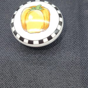 May include: A white ceramic cabinet knob with a black and white checkered border. The center of the knob features a yellow bell pepper with green leaves.