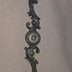 May include: Ornate, antique-style cabinet pull with a dark bronze finish. The decorative handle features scrolling leaf and floral designs, with a central circular element. A vintage-inspired hardware piece.
