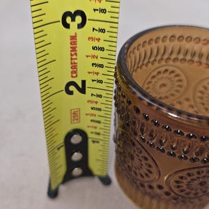 May include: Amber-colored glass votive candle holder with a decorative pattern. The candle holder is approximately 3 inches tall. A yellow Craftsman tape measure is next to the candle holder.