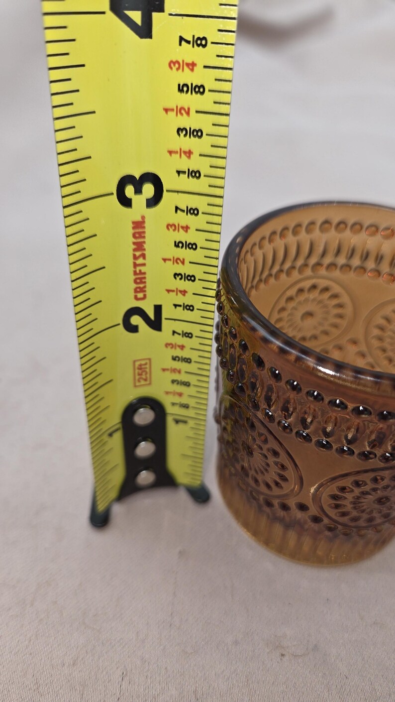 May include: Amber-colored glass votive candle holder with a decorative pattern. The candle holder is approximately 3 inches tall. A yellow Craftsman tape measure is next to the candle holder.