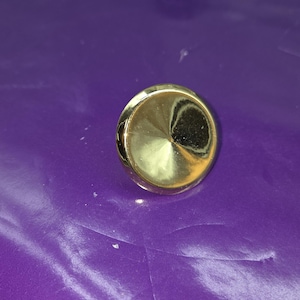 May include: A gold-colored, circular button with a conical center. The button is set against a purple background. The button's surface reflects light, creating highlights and shadows.