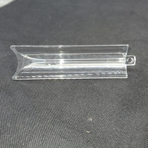 May include: A clear, rectangular plastic item with a curved end and a small circular hole on the opposite end. The item has a series of parallel lines running along its length. The object is on a black surface.
