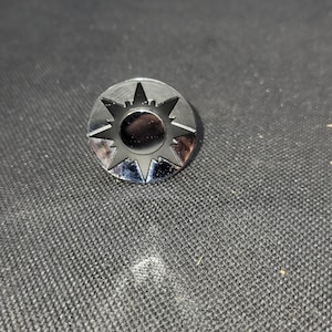 May include: A shiny, silver-colored metal pin with a sunburst design. The pin features a central circle surrounded by pointed rays, creating a star-like appearance. The pin is circular and has a reflective surface.